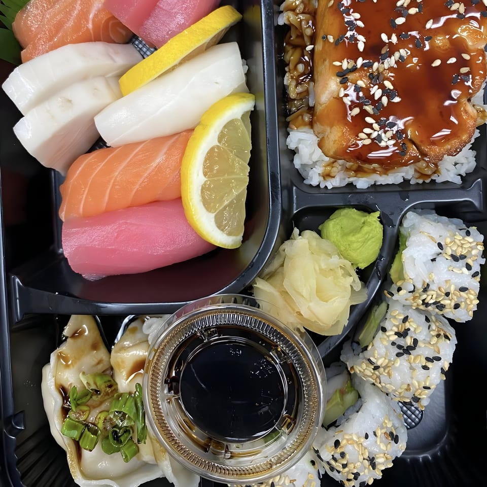 A bento box filled with sushi, sashimi, dumplings, and a salmon teriyaki rice bowl.