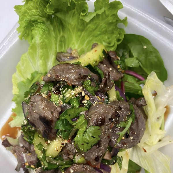 Thai beef salad with lettuce, cilantro, cucumbers, and a savory dressing in a white container.
