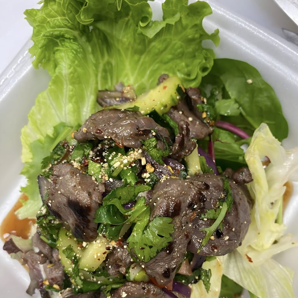 Thai beef salad with lettuce, cilantro, cucumbers, and a savory dressing in a white container.