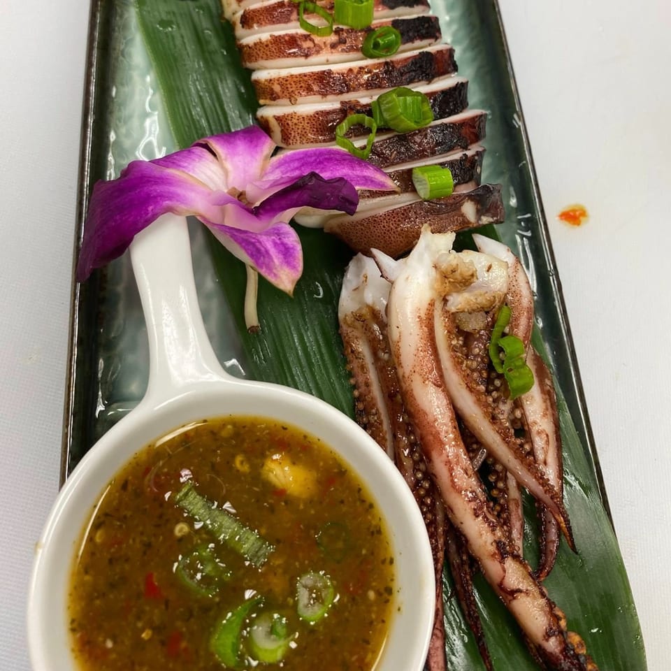 Grilled squid, sliced and whole, with a side of dipping sauce and a purple orchid.